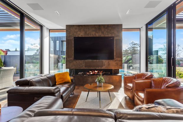 Bright contemporary living room with extensive floor-to-ceiling glazing, a central dark stone or textured feature wall containing a television and fireplace, and leather lounge seating arranged around a round coffee table. The room opens to an outdoor terrace and landscaped surroundings.