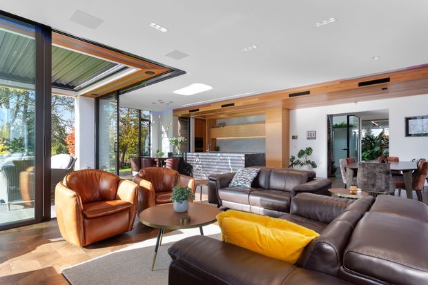 A bright, open-plan contemporary living room with extensive floor-to-ceiling glazing, leather lounge seating, a timber coffee table, and a visible dining area. Warm timber wall panelling and built-in joinery frame a kitchen or bar area at the rear, while a large glazed opening connects the room to the outdoors.