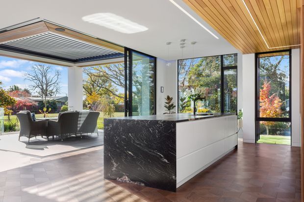 Bright contemporary kitchen with a large island featuring a dramatic dark veined stone waterfall end and white flat-panel cabinetry. The room has a warm timber slatted ceiling, parquet-style timber flooring, extensive black-framed floor-to-ceiling glazing, and direct connection to a covered outdoor entertaining area and landscaped garden.