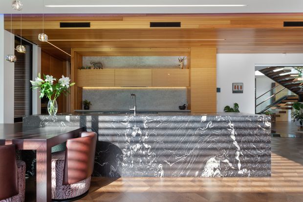 A sophisticated open-plan kitchen dominated by a large waterfall island clad in dramatic veined dark stone. Warm timber cabinetry and a timber-lined ceiling create a cohesive feature wall, with integrated appliances, open shelving, pendant lighting, and a dining area in the foreground. A stairway and adjoining living space are visible beyond the kitchen.