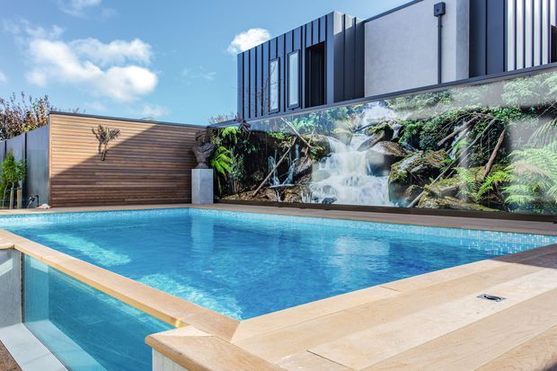 A contemporary residential outdoor pool courtyard with a large rectangular swimming pool, pale stone coping and paving, a timber-clad privacy wall, and a dramatic planted waterfall feature. The modern two-storey building behind has dark vertical cladding, rendered walls, and expansive glazing. Bright daylight and lush planting create a resort-like atmosphere.