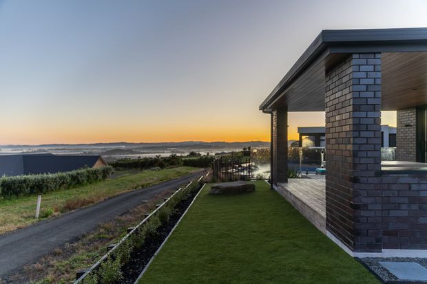 Exterior view of a contemporary single-storey house at sunset, with dark brick piers, a broad flat roof, timber-lined soffits, and expansive outdoor living areas. The landscaped lawn and paved terrace overlook a coastal or rural landscape, with a swimming pool visible beside the covered veranda.