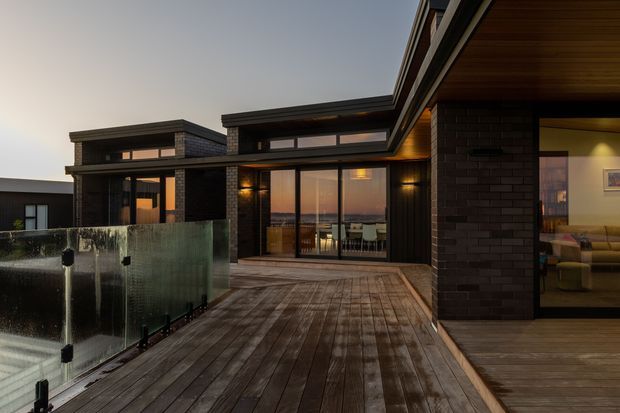 Twilight exterior view of a contemporary, low-profile residence arranged around a broad timber deck. Dark brick walls, expansive glazed sliding doors, warm timber soffits, and integrated exterior lighting create a sheltered outdoor living area. A glass balustrade borders the deck, while a living room is visible through the glazing on the right and dining furniture is visible inside at the centre.