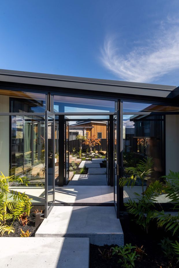 A bright exterior view through a series of black-framed glazed doors and covered openings into a landscaped courtyard. Concrete stepping paths lead between low modern buildings, with timber-clad elements, lush planting, and strong sunlight beneath a clear blue sky.