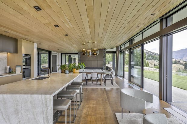 Open-plan contemporary kitchen, dining and living space with a large stone island, timber cabinetry, integrated appliances, dining table and lounge seating. The room has extensive floor-to-ceiling glazing with views to a mountainous landscape, and a distinctive warm timber-lined ceiling with recessed lighting.