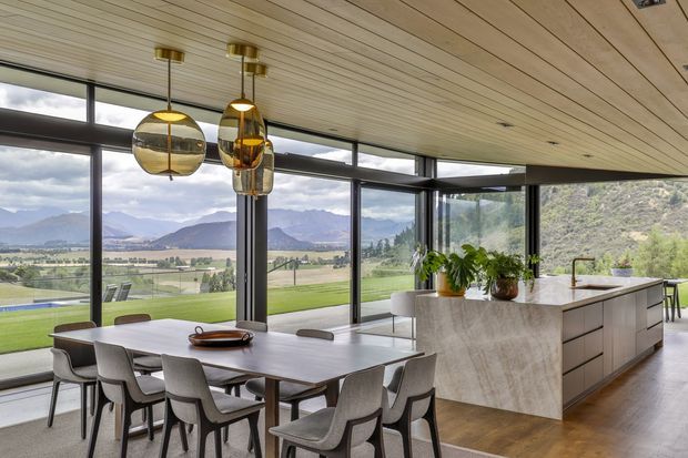Contemporary open-plan kitchen and dining area with a large stone island, timber-lined ceiling, dark-framed floor-to-ceiling glazing, pendant lights, and a dining table overlooking an expansive mountain and valley landscape.