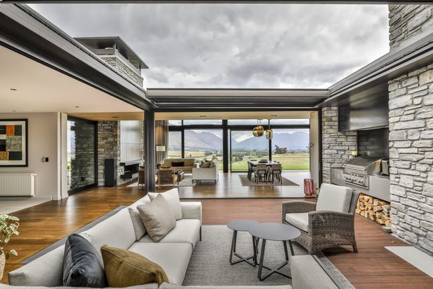 Open-plan contemporary living and dining area with a large sectional sofa, armchairs, layered coffee tables, timber flooring, and extensive glazing overlooking a mountainous landscape. Exposed stone walls frame the room, while a built-in outdoor-style kitchen or barbecue zone with stacked firewood sits to the right. The space has a partially vaulted or high ceiling and strong indoor–outdoor connectivity.