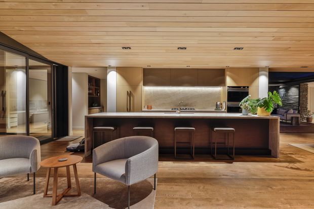 Warm, open-plan contemporary kitchen with a large dark timber island and integrated bar seating, set beneath a full timber-lined ceiling. The space includes light wood flooring, dark cabinetry, stone-look benchtops and splashback, integrated appliances, and a small adjoining lounge area with upholstered armchairs. Floor-to-ceiling glazing and dark-framed doors are visible at the sides.