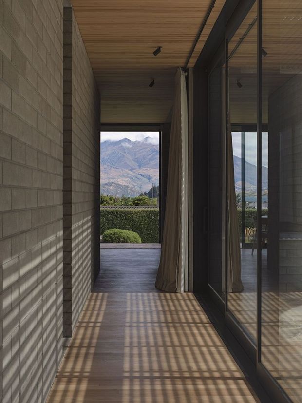A narrow, minimalist interior passage with exposed grey masonry block walls, a warm timber-lined ceiling, and dark timber flooring. Full-height black-framed glazing and reflective glass run along the right side, with curtains gathered near the opening. Strong patterned sunlight and shadows fall across the floor and walls. The passage opens toward a landscaped exterior and a dramatic mountain landscape visible beyond.