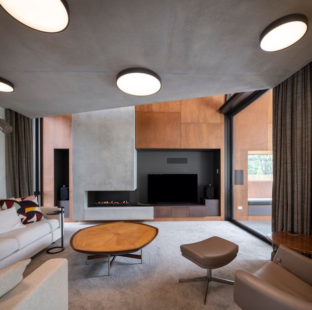 A sophisticated contemporary living room with a low concrete ceiling, large round ceiling lights, full-height glazing, and dark curtains. The room features a built-in timber and dark grey entertainment wall with a linear fireplace, television, and integrated cabinetry. A light sectional sofa, sculptural patterned cushions, round timber coffee table, and leather lounge seating create a warm, refined setting. An outdoor terrace is visible through the glazing.