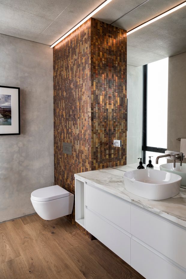 Contemporary bathroom featuring a wall-mounted toilet, white floating vanity with integrated drawers, marble-look benchtop, vessel basins, and dark metal fixtures. A full-height wall clad in small vertically arranged timber-toned tiles forms the main feature, contrasted with smooth grey walls, warm timber flooring, large black-framed window, and linear recessed ceiling lighting.