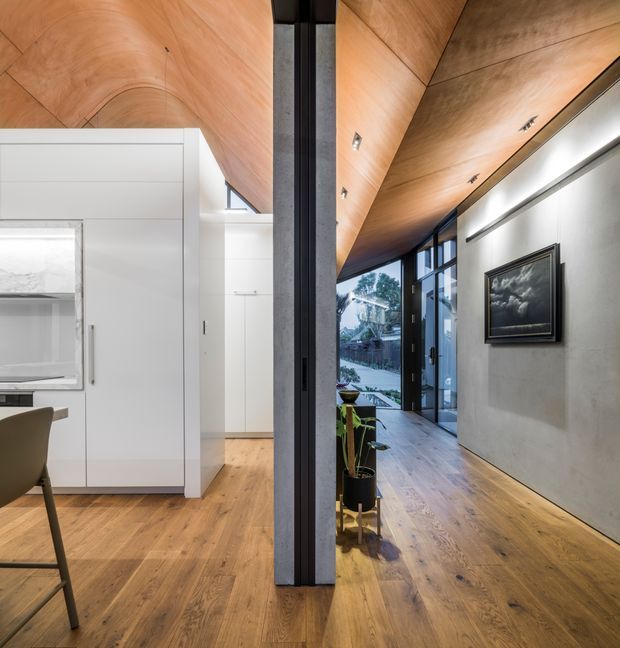 Interior view of a contemporary open-plan residential space with a warm timber-lined angled ceiling and matching timber flooring. A white built-in kitchen or storage volume occupies the left side, while a long concrete-finished wall with framed artwork and linear lighting extends along the right. Black-framed full-height glazing opens toward an exterior garden, and a dark structural column divides the spaces.