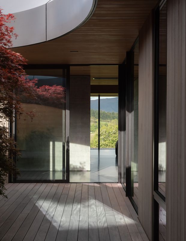 A refined covered architectural passage or terrace with full-height dark-framed glazing, timber-lined ceiling, vertical timber wall cladding, and wide timber decking. The space opens toward a green, mountainous landscape, while sculptural curved white roof forms and reflected Japanese maple foliage add visual interest. The composition is minimalist and strongly connected to the outdoors.