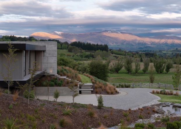 A contemporary low-profile house set within a landscaped rural valley, viewed from outside. The building has a dark flat roof, pale grey horizontal cladding or concrete-like wall surfaces, and large glazed openings with vertical timber-toned screening. A gravel driveway and stepped path lead through drought-tolerant planting toward the house, with open pasture, trees, and snow-dusted mountains beyond.