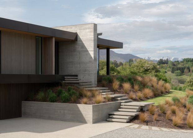 Contemporary low-profile concrete residence set into a landscaped, sloping site. The home features broad dark roof planes, exposed board-formed concrete walls, vertical timber-toned cladding, deep overhangs, and a series of stepped concrete paths and terraces. Ornamental grasses and native-style planting frame views toward a green valley and distant snow-capped mountains.