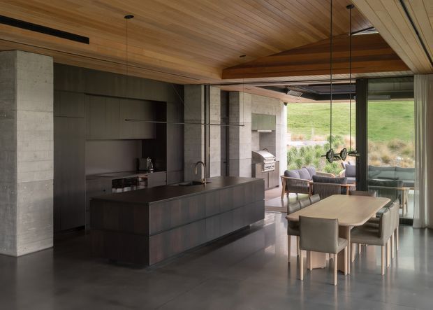 A sophisticated open-plan kitchen and dining area with a large dark island, integrated cabinetry, concrete structural walls, and a warm timber-lined ceiling. Full-height glazing opens to a furnished outdoor terrace and grassy landscape, while a dining table and living seating occupy the adjacent space. The interior has a refined contemporary feel with strong indoor-outdoor connection and natural materials.