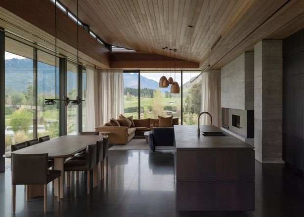Open-plan contemporary kitchen, dining, and living space with extensive floor-to-ceiling glazing overlooking a mountainous rural landscape. The room features a large dark kitchen island, integrated sink and tap, timber-lined ceiling and walls, polished dark flooring, a concrete fireplace wall, dining table, lounge seating, and pendant lighting. The design has a warm, refined natural-material palette and strong indoor-outdoor visual connection.