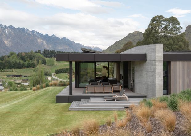 Contemporary single-storey residence set in a landscaped alpine setting, with a strong connection between the interior and an outdoor terrace. The building combines dark horizontal cladding and framing with a large exposed concrete wall, extensive glazing, a low-pitched roof, and broad timber decking. Mountain peaks, manicured lawn, native-style grasses, and mature trees surround the house.