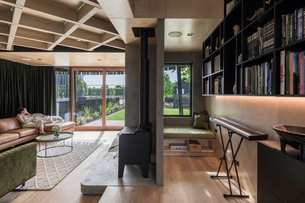 A refined open-plan living room with a warm timber interior, black built-in shelving, and a compact black freestanding wood stove dividing the space. The room includes a sofa and lounge seating, a rug and coffee tables, a built-in window seat, an electronic keyboard, and full-height glazed doors opening to a landscaped outdoor area. The ceiling features a distinctive exposed timber grid with recessed lighting, while integrated shelving has warm accent lighting.