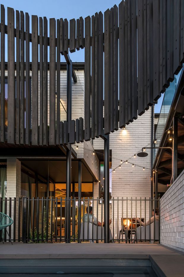 A contemporary multi-level courtyard or outdoor entertaining area enclosed by dark vertical timber screening and metal railings. White brick walls, exposed steel posts, warm festoon lighting, outdoor seating, and a visible fire feature create a sheltered architectural courtyard beside a partially visible pool or water feature.