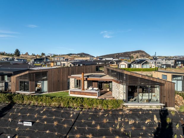A contemporary single-storey residential house viewed from above, featuring dark timber cladding, low-pitched roof forms, extensive glazing, projecting horizontal slatted screens, and a central outdoor courtyard or terrace with a built-in fireplace or chimney element. The surrounding development and dry hills are visible in the background.