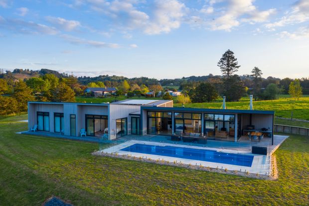 A low, single-storey contemporary home set in a spacious rural landscape. The building has a flat roof, rectilinear modular form, extensive floor-to-ceiling glazing, dark-framed windows, and pale blue-grey exterior wall panels. A long rectangular swimming pool and paved terrace run along the glazed living areas, with open lawns and mature trees surrounding the property.