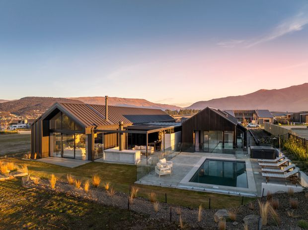 Exterior view of a contemporary single-storey residence in a mountainous setting, composed of several dark-clad gabled volumes with extensive floor-to-ceiling glazing. The home opens onto a paved terrace with outdoor seating and a rectangular swimming pool, surrounded by landscaped grasses and a low fence. Warm sunset light highlights the timber-toned interiors and exterior elements.