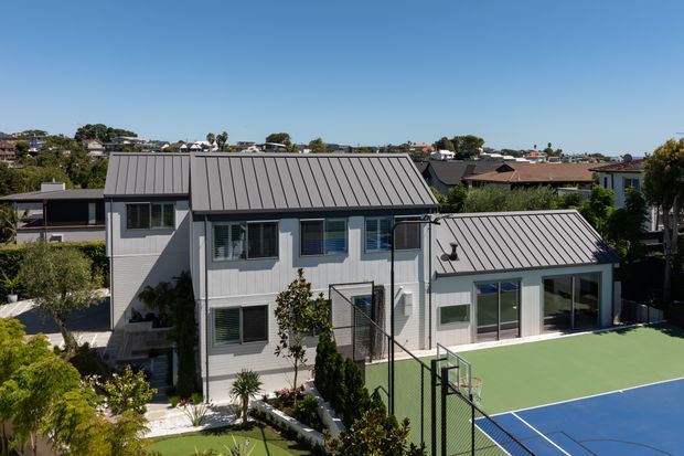 A contemporary multi-level residential home viewed from above, with light grey cladding, dark grey standing-seam metal roofing, and extensive glazed doors and windows. The property includes a prominent green and blue outdoor tennis or sports court in the foreground, landscaped planting, and a dense suburban neighbourhood beyond.