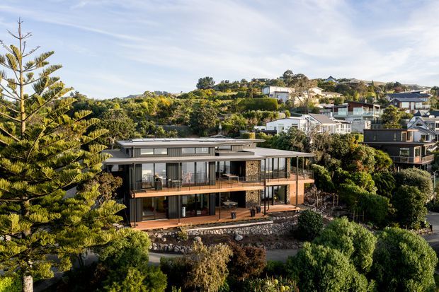 Exterior aerial or elevated view of a contemporary multi-level residential house set on a densely planted, sloping suburban site. The home features broad glazing, dark-framed windows, timber-toned balcony decking and wall accents, a low-pitched roof, and prominent cantilevered outdoor living areas. Other hillside homes and mature coastal-looking vegetation surround the property.