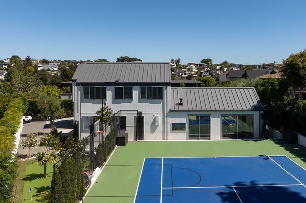 Exterior view of a contemporary two-storey light-coloured home with dark standing-seam metal gabled roofs, large glazed openings, and a full-size blue-and-green tennis court in the foreground. The house sits within a suburban neighbourhood and landscaped grounds.