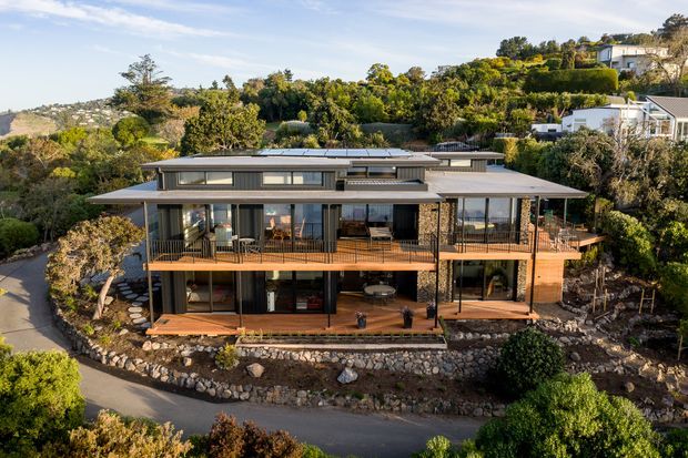 A wide aerial exterior view of a contemporary multi-level home set into a landscaped hillside. The house features broad flat roof planes, extensive glazing, timber decks and balconies, dark-framed windows, exposed structural columns, and stone retaining walls. Dense greenery surrounds the property, with a winding driveway in the foreground and neighbouring hillside homes visible in the distance.
