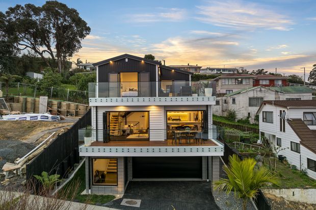 A contemporary multi-level hillside residence viewed from outside at dusk. The home has dark vertical cladding on the upper level, light horizontal cladding below, broad glazed openings, two projecting balconies with glass balustrades, warm interior lighting, and an integrated garage at ground level. Neighbouring houses and landscaped garden areas surround the property.