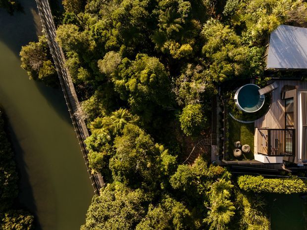 Aerial view of a compact building and timber deck nestled within dense lush vegetation beside a narrow calm waterway. The property includes a small circular plunge pool, outdoor seating, planted garden areas, and a light metal roof, with a narrow edge walkway or retaining structure running along the water.