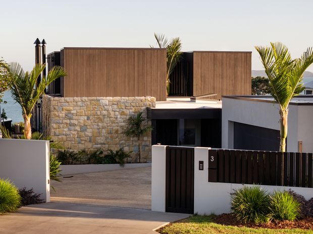 Contemporary multi-level residential facade viewed from the street, featuring warm vertical timber-look cladding, a prominent pale natural-stone feature wall, dark metal-framed openings and garage door, white rendered walls, and a gated driveway. Palm trees and drought-tolerant landscaping frame the property.