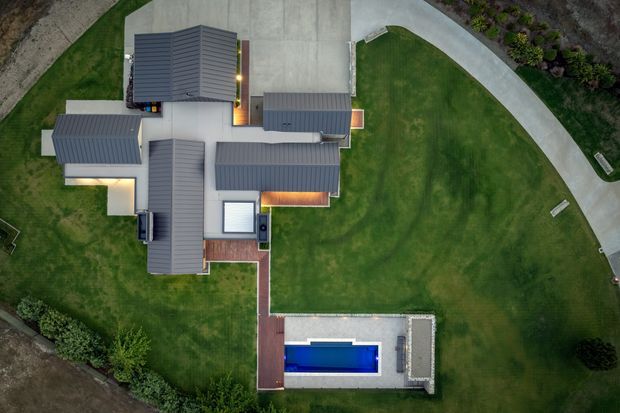Aerial top-down view of a large contemporary residential property with multiple dark grey metal-clad roof volumes, concrete pathways and drive areas, expansive manicured lawns, timber decking, and a rectangular swimming pool.