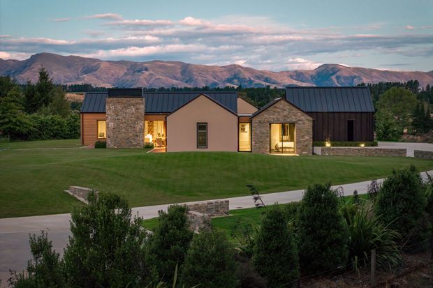 A low, contemporary single-storey residence set within a broad landscaped lawn, with several connected gabled volumes, dark metal pitched roofs, warm stone-clad walls, and muted rendered sections. Warm interior lighting glows through the windows and recessed entry areas. A paved driveway or walkway crosses the foreground, while a dramatic mountain range forms the distant backdrop.