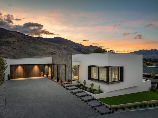 Exterior view of a contemporary single-storey residence at sunset, set against rugged mountains. The home features crisp white rendered walls, a low flat roofline, extensive dark-framed glazing, a stone-clad entry volume, and a warm timber garage door. A broad paved driveway, stepping-stone path, landscaped planting, and a neatly edged lawn surround the house.