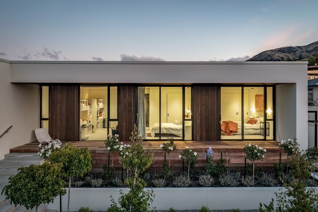A low, modern single-storey residence viewed from the landscaped front garden at dusk. The white flat-roofed facade features extensive floor-to-ceiling glazing, dark timber privacy screens, warm interior lighting, and a raised timber terrace with planted garden beds. Bedrooms and interior furnishings are visible through the glass, with a mountainous backdrop beyond.