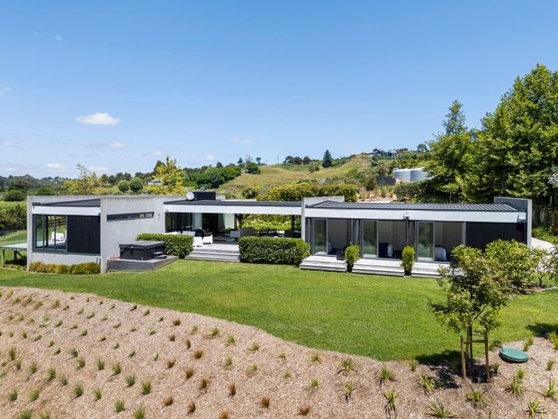 Wide exterior view of a low, single-storey contemporary residence set in a landscaped rural or semi-rural site. The house has a strongly horizontal composition with flat rooflines, dark-framed glazing, pale rendered walls, covered outdoor areas, manicured lawn, planted garden beds, and rolling green landscape beyond.