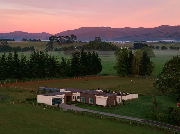 Aerial view of a low, modern rural residence set within expansive green farmland, with mature trees, a long driveway, distant mountain ranges, and a pink sunset sky. The home has a flat-roofed, horizontally composed form with pale rendered walls and dark-framed glazing.