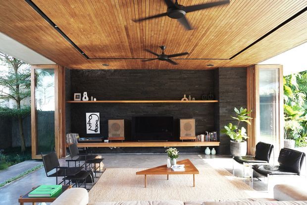 A bright, open-plan living room with a dramatic dark brick feature wall and built-in timber shelving surrounding a television. The room has a warm timber slatted ceiling with two black ceiling fans, extensive glazed openings and open glass doors to lush greenery, black leather lounge chairs, a pale sofa, a low timber coffee table, and a large light rug. The setting has a refined mid-century-inspired indoor-outdoor character with strong natural light.