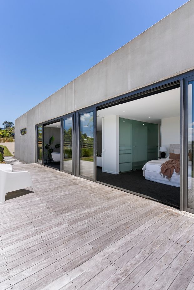 Exterior architectural view of a minimalist single-storey concrete residence opening onto a broad timber deck. Full-height dark-framed sliding glass doors reveal a calm bedroom interior with a bed, dark carpet, white walls, and a pale green glazed or tiled feature wall. The deck and glazing create a strong indoor-outdoor connection under a clear blue sky.