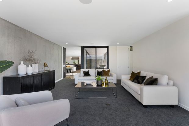 Bright contemporary living room with white sofas and armchairs arranged around a low black metal-and-glass coffee table. The room features dark grey carpet, a large grey textured feature wall, white walls and ceiling, black-framed glazed internal doors, and minimalist built-in cabinetry with sculptural decor. A dining or kitchen area is visible beyond the glazing and opening.