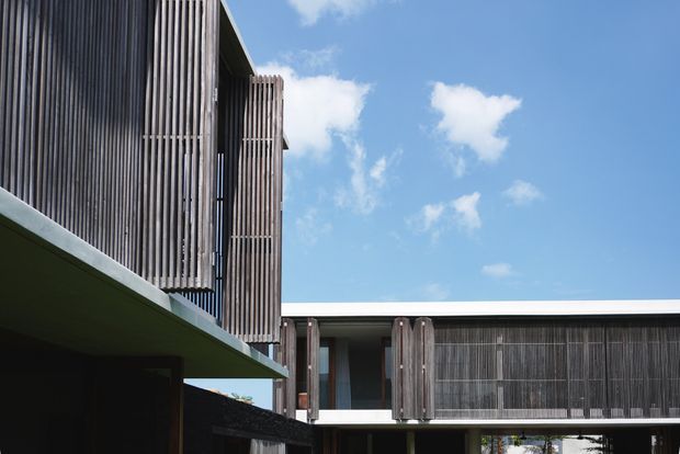 Exterior architectural view of a contemporary multi-level building with strong horizontal and vertical composition. Dark timber or metal-look vertical screening wraps the upper facade and conceals glazing, while broad flat roof planes and projecting cantilevered forms create a sculptural, rectilinear appearance. Bright blue sky is visible around the structure.