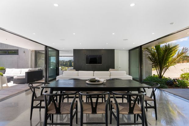 A bright, contemporary open-plan living and dining area with a long dark dining table and black-framed chairs in the foreground. A white sectional sofa faces a wall-mounted television set into a dark vertical feature wall, while expansive glazed openings connect the room to a landscaped outdoor courtyard or terrace. The space has a polished concrete floor, white ceiling with recessed downlights, and strong indoor-outdoor flow.