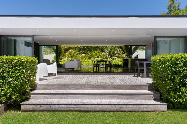 Exterior view of a sleek single-storey modern home opening onto a broad timber deck. Large glazed openings connect the interior living and dining areas to the deck, with outdoor seating and dining furniture framed by neatly clipped hedges and landscaped greenery. Wide timber steps lead up from the lawn, and a thin flat roof projects over the terrace.
