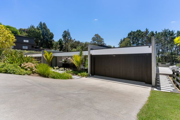 A contemporary single-storey residence viewed from the street, with a broad concrete driveway leading to a dark slatted double garage door. The house has crisp white and charcoal geometric volumes, low-pitched rooflines, timber-look cladding, and landscaped planting with palms and ornamental grasses. A stepped walkway and dark horizontal fencing extend along the right side, with mature trees in the background under a clear blue sky.