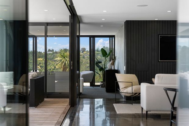 A refined contemporary living room with polished concrete flooring, white upholstered seating, and a black vertically panelled feature wall with a wall-mounted television. Floor-to-ceiling glazing and dark-framed sliding doors connect the room to a leafy outdoor setting, while a glass-partitioned space is visible to the left.