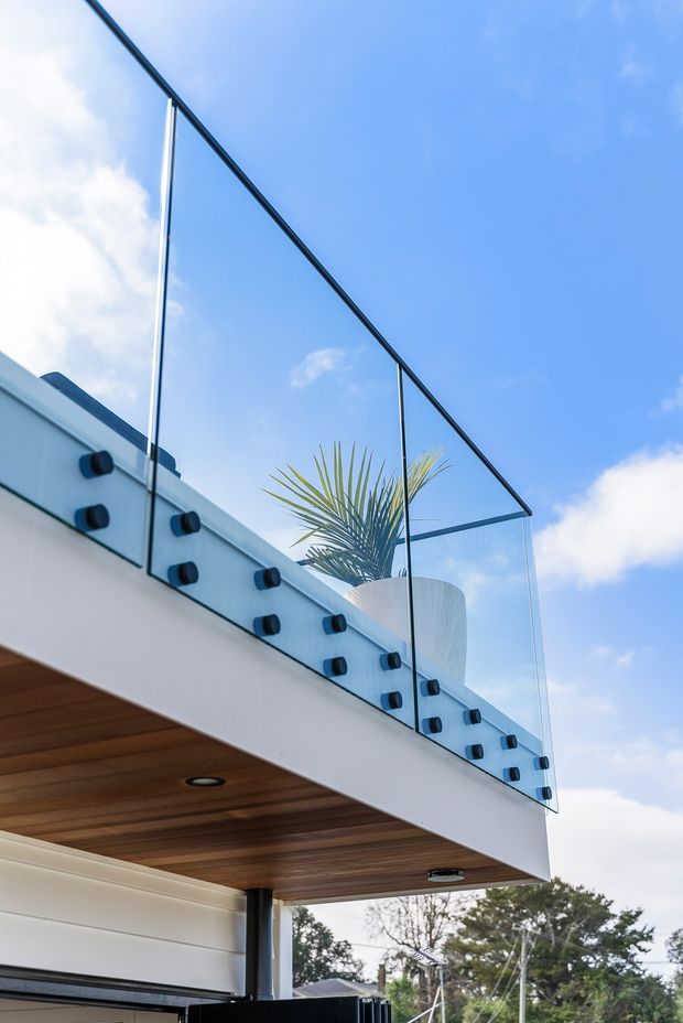 Low-angle exterior architectural detail showing a contemporary upper-level balcony or terrace with clear frameless glass balustrading, dark fixings, and a white structural edge. The underside features warm timber-lined soffit boards and recessed lighting, with a potted palm visible behind the railing. Blue sky and surrounding trees form the background.