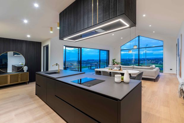 A spacious open-plan contemporary kitchen and living area with a large dark island, integrated cooktop, dramatic overhead rangehood enclosure, black vertical wall panelling, light timber flooring, and expansive blue-tinted glazing overlooking a distant landscape. A dining table and living seating are visible beyond the kitchen.